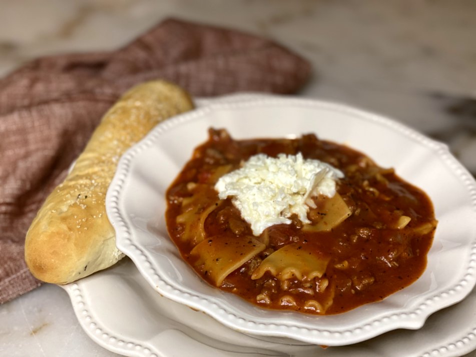 hearty lasagna soup in a white dish with red napkins, cheese on top and a garlic breadstick on the side. 