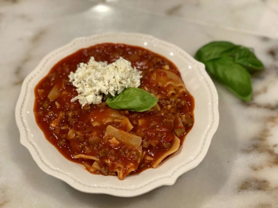 hearty lasagna soup in a white dish with red napkins, cheese on top and a garlic breadstick on the side. with basil leaves on top and to the side.