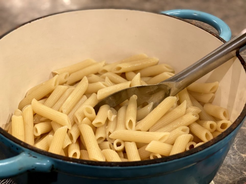 cooked penne pasta in a large blue dutch oven with a steel slated spoon.