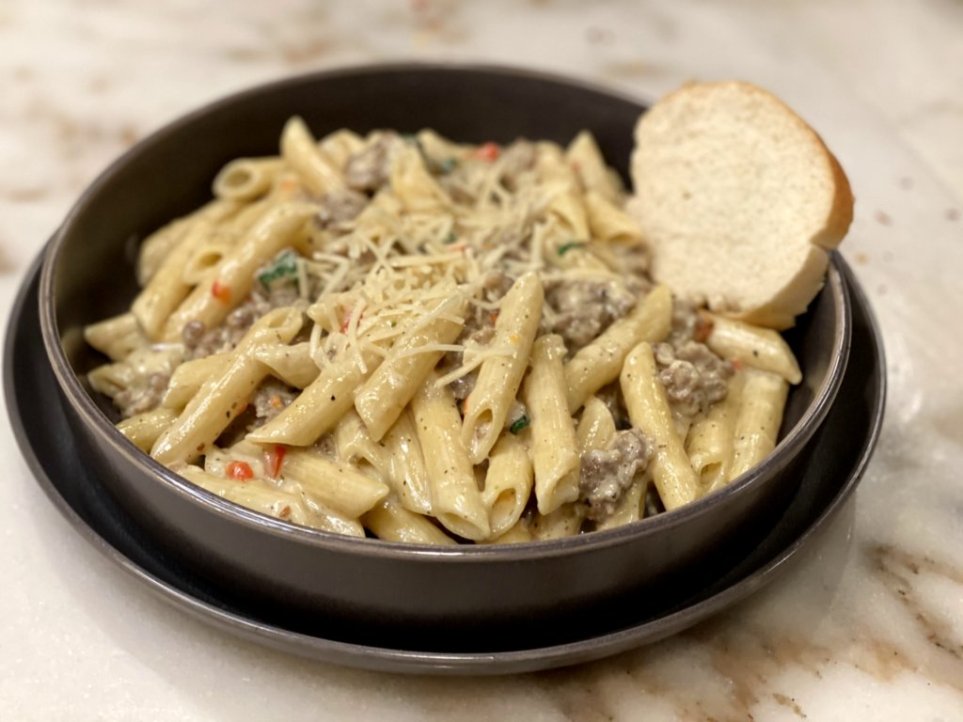 creamy italian sausage pasta served in a black bowl and plate with a slice of bread and sprinkled with Parmesan cheese.