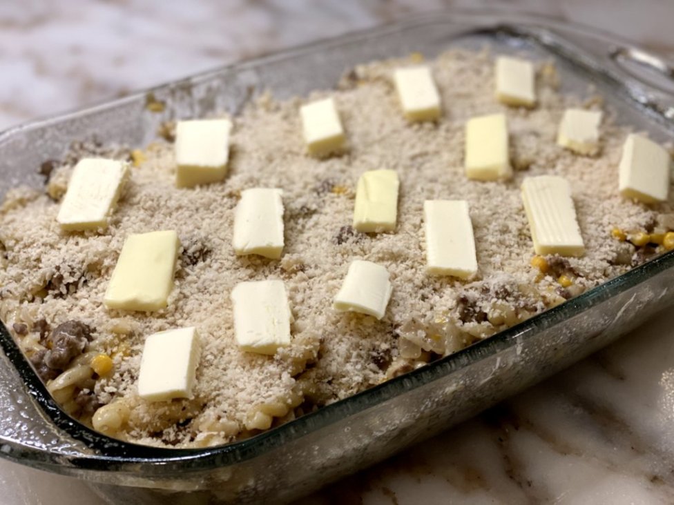 ground beef with noodles, corn, sour cream, cream of chicken soup with bread crumbs browned on top in a glass casserole dish with slices of butter on top.