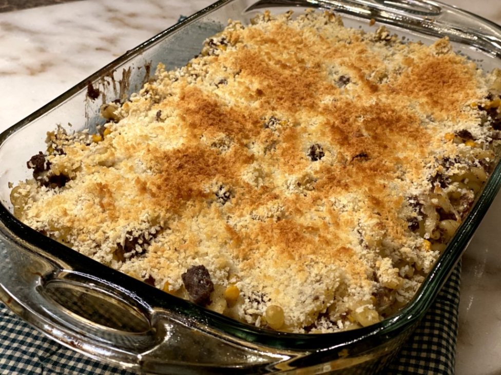 hamburger noodle bake with cooked ground beef with corn, sour cream, milk, cream of chicken soup and pasta noodles on the side in a glass baking dish on a green and white hot pad