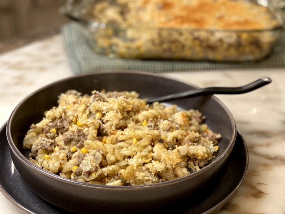 cooked ground beef with corn, sour cream, milk, cream of chicken soup and pasta noodles on the side in a glass baking dish on a green and white hot pad