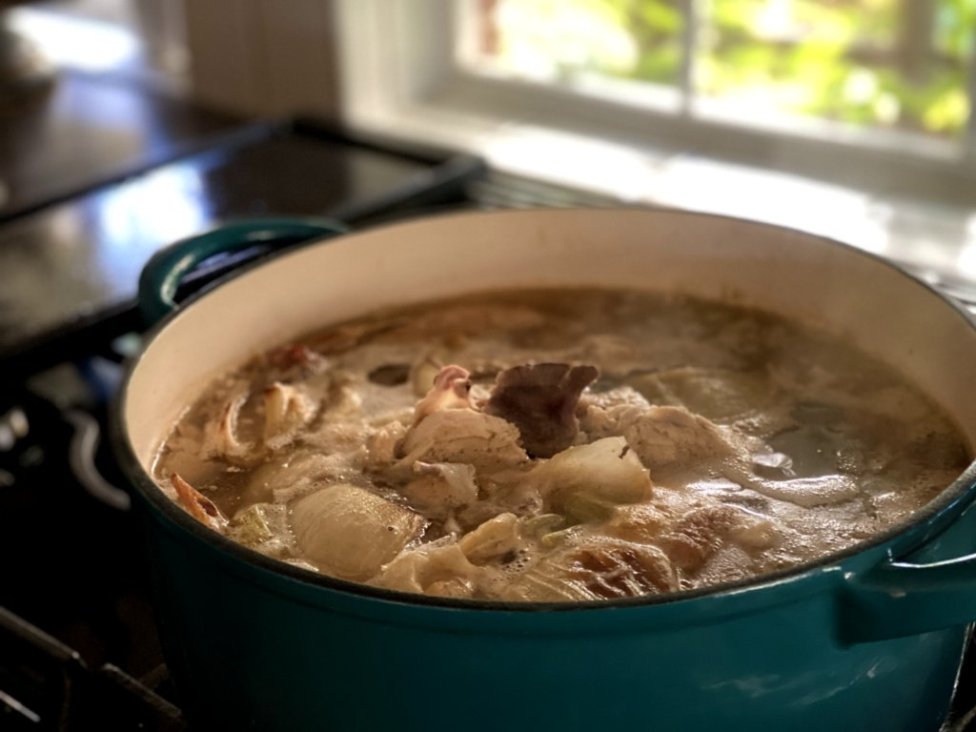 Scratch roasted homemade chicken stock and broth simmering over the stove in a blue dutch oven pot.