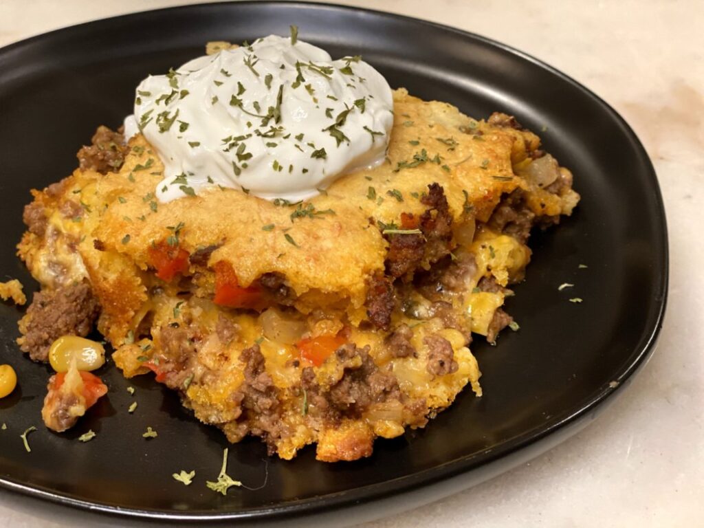 Tamale pie casserole filled with ground beef, peppers, onions, corn muffin mix, and topped with sour cream on a black plate. 