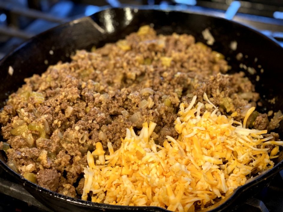 adding shredded cheddar cheese to the ground beef and sauce in a black skillet