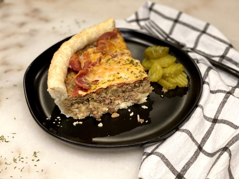 one slice of bacon cheeseburger pie on a black plate with a black fork with dill pickle slices on the side. 