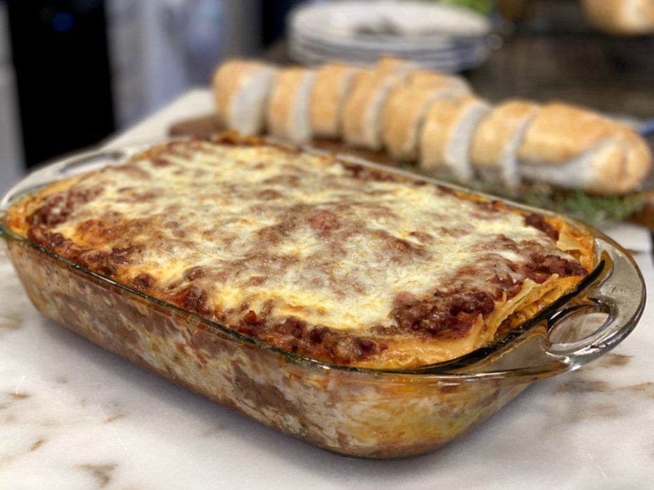 lasagna in a clear glass casserole dish with bread and plates in the background