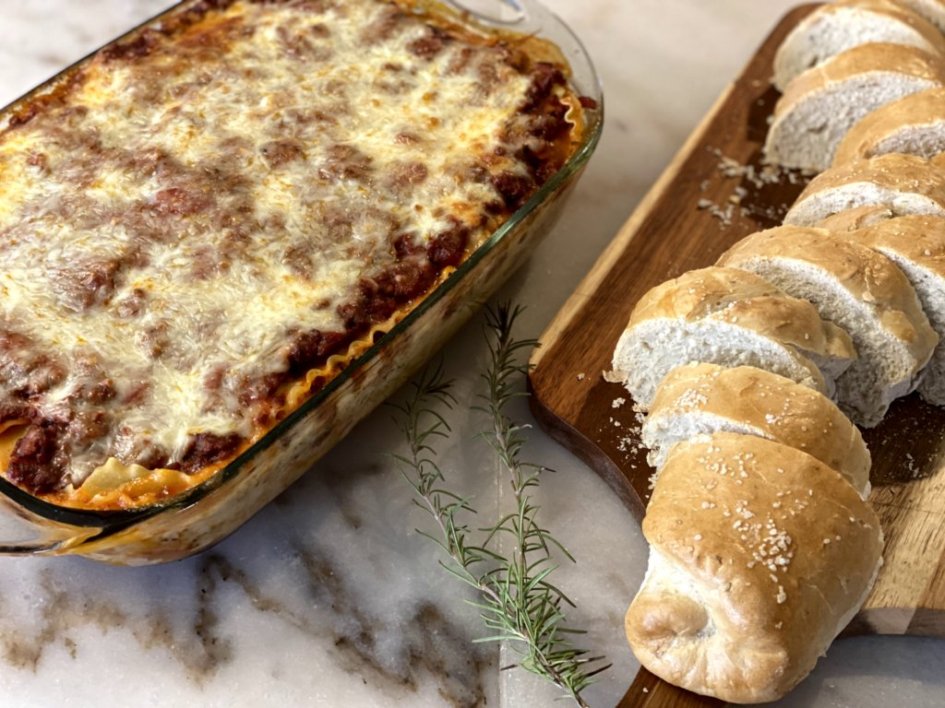 coogan's kitchen lasagna recipe made from scratch with rosemary french bread on the side on a wood cutting board.