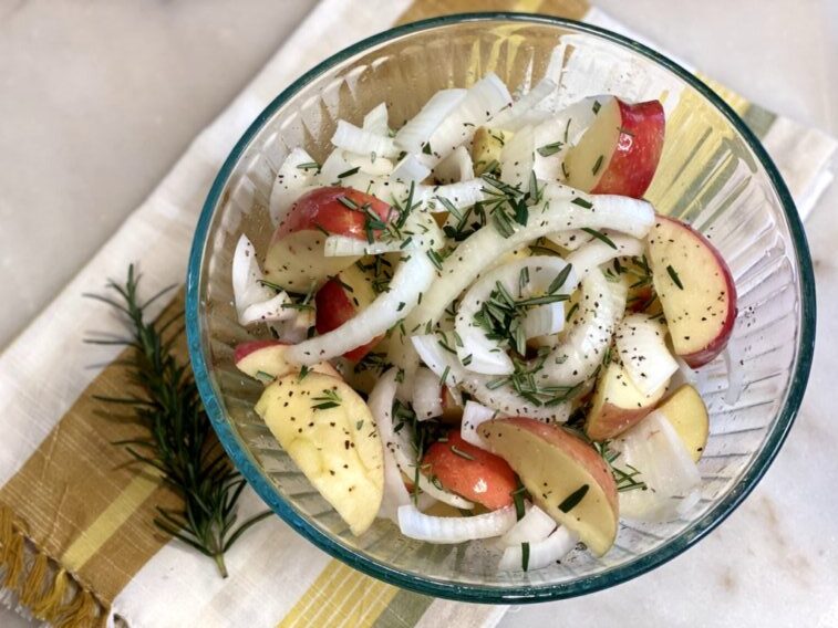 sliced apples, onions, chopped rosemary, and seasonings tossed in a glass bowl with olive oil. 