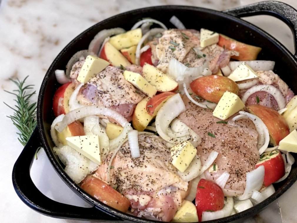 Raw chicken, vegetables, and apples placed in a black cast iron skillet and topped with wedges of butter before placed in the oven. 