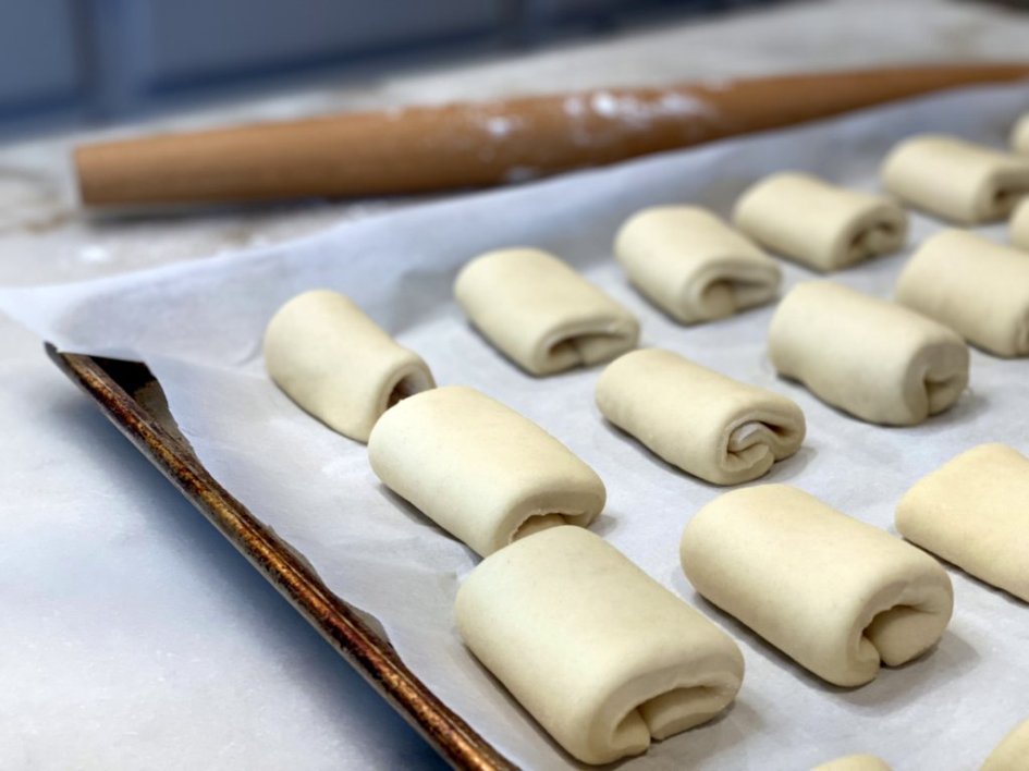 preparing the dinner rolls for baking - dough is folded on a large baking tray with parchment paper with a floured rolling pin in the background. 