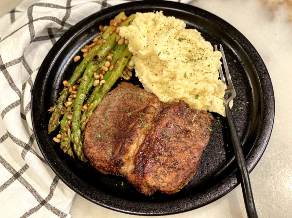 Easy asparagus with pine nuts, grilled pork chops and cheddar garlic mashed potatoes on a black plate with a fork and napkin.