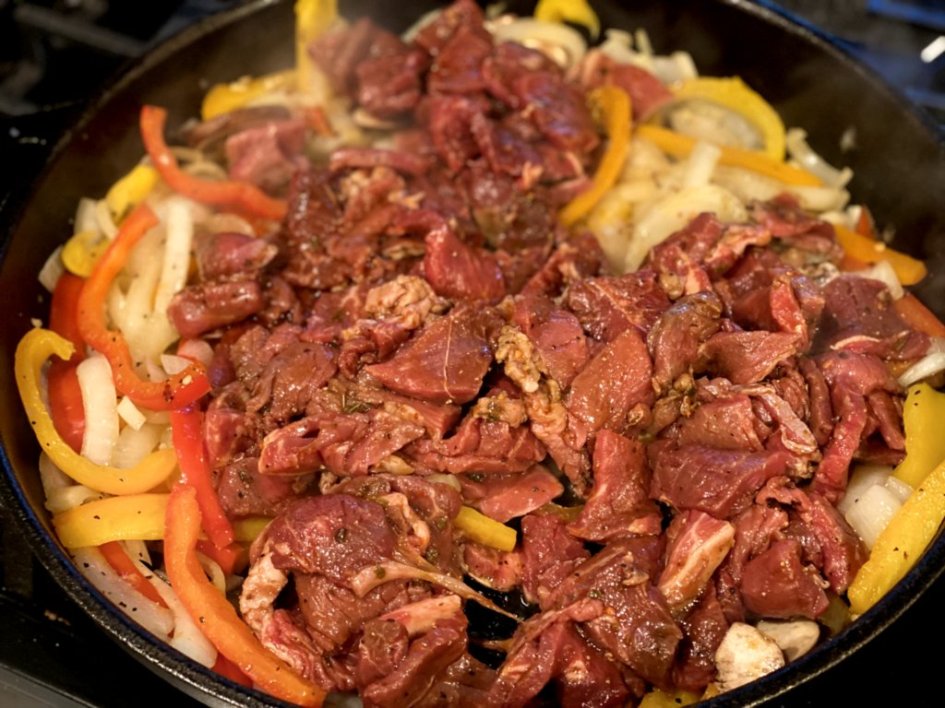 Adding the marinated steak to the skillet of softened peppers and onions. 