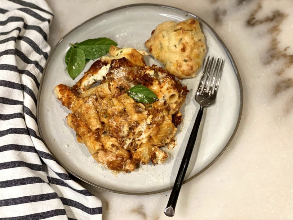 Creamy Baked Pasta served on a white plate with a rosemary cheddar drop biscuit with garlic butter and basil leaves as garnishment.