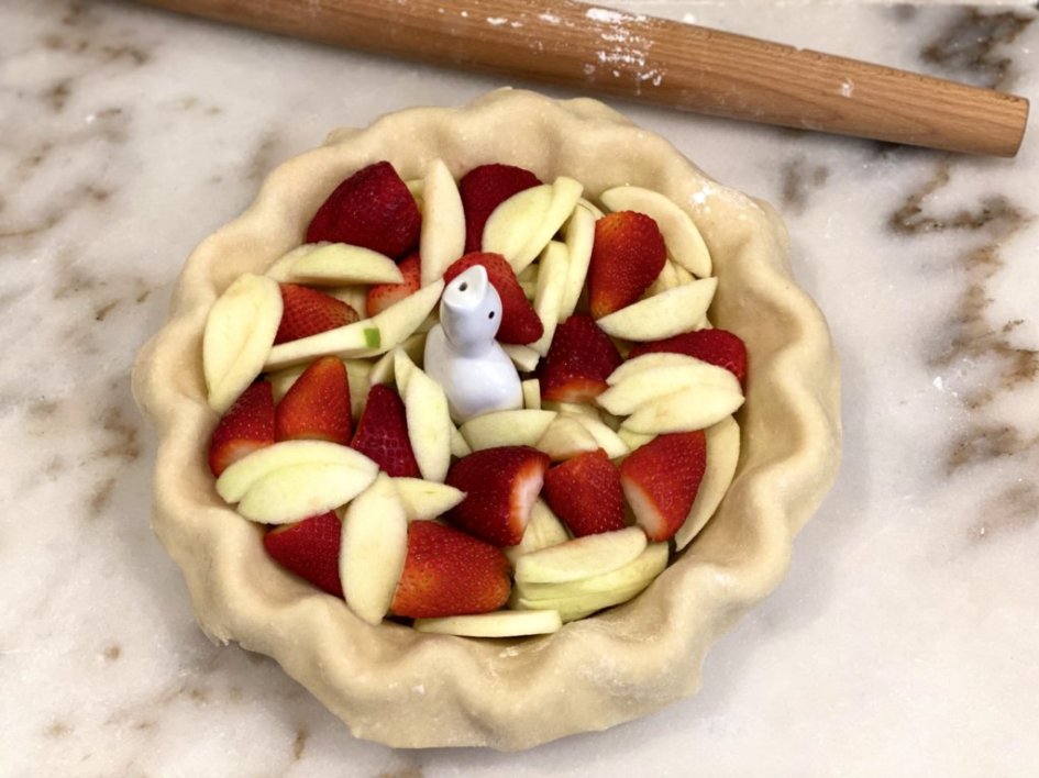 pie crust with freshly cut granny smith apples and sliced strawberries with a ceramic pie bird in the middle. rolling pin in the background. 