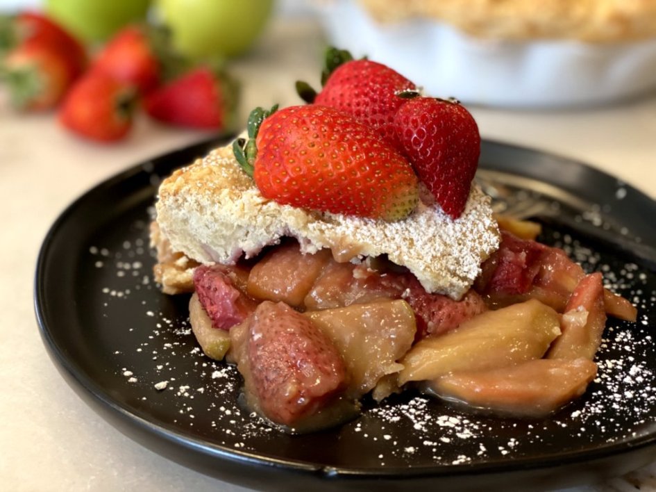 Delicious apple strawberry pie with powdered sugar and fresh strawberries on top. Sliced and sitting on a black plate. cooganskitchen.com