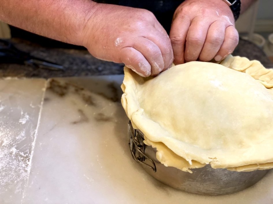 Adding pastry dough to the top of the timballo in the shape of a little dome and then crimping the edges. 