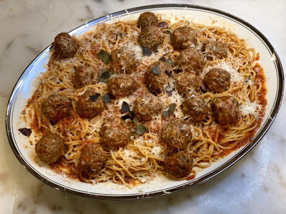 family style plate of juicy meatballs with spaghetti noodles, homemade tomato sauce, and basil leaves on a large white serving platter. 