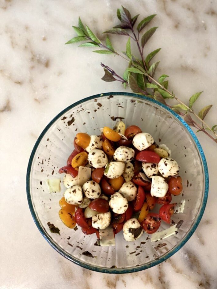 a glass bowl of colorful cherry tomatoes, fresh mozzarella pearls, shredded parmesan cheese, fresh basil leaves that's been tossed gently in olive oil and balsamic vinegar. 
