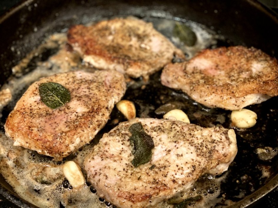 pork chops frying in a skillet with sage leave, garlic, butter, and olive oil. 