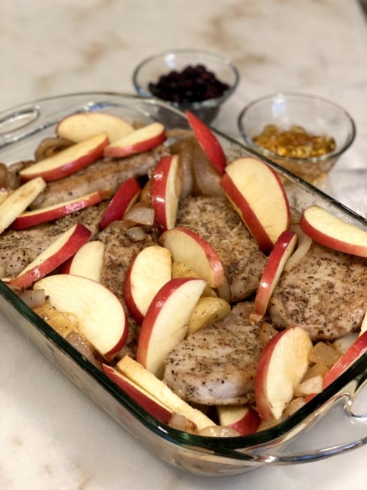 Sliced apples, potatoes, sweet onions, and seared pork chops ready to bake in the oven in a glass baking dish. 