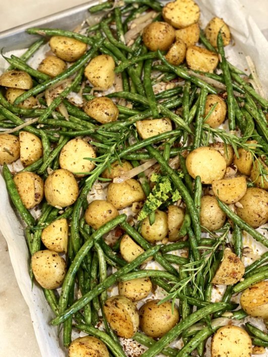 green beans and chopped gold potatoes on a baking sheet seasoned with salt and pepper and springs of rosemary and thyme